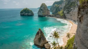 Pemandangan Pantai Nusa Penida di Bali dengan air laut biru jernih, pasir putih, pohon kelapa, dan formasi batu karang yang indah.