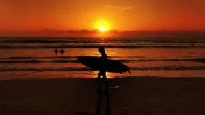 Peselancar menikmati matahari terbenam yang memukau di Pantai Kuta, Bali dengan langit berwarna oranye.